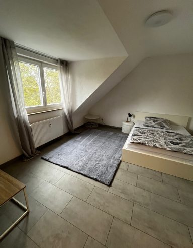 Helles Schlafzimmer mit Schrägwand, Bett, Teppich und Fenster mit Vorhängen.