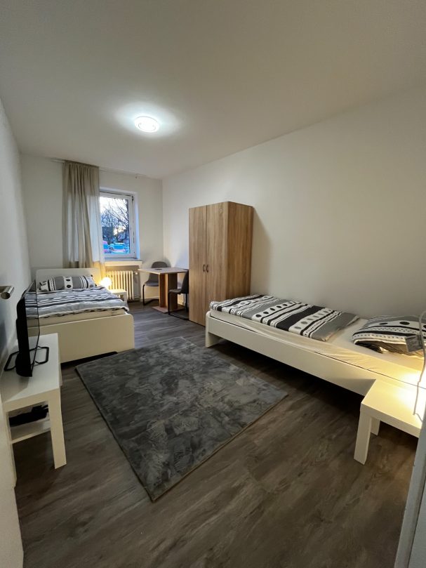 Helles Zimmer mit zwei Betten, Tisch, Stuhl und einem Schrank. Holzfußboden und Teppich.