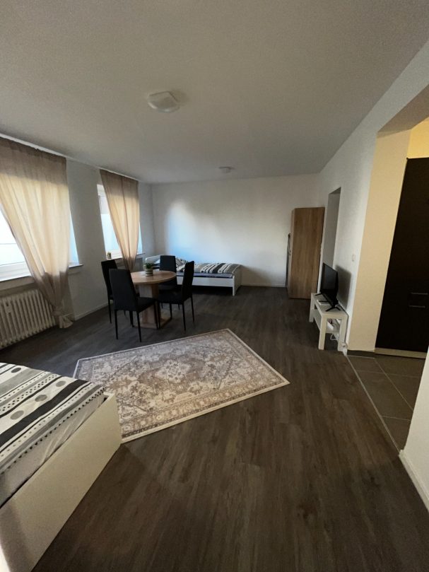 Helles Wohnzimmer mit Esstisch, Stühlen, Sofa und TV. Fenster mit Vorhängen.