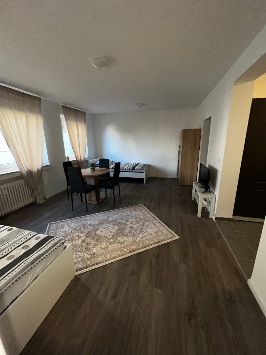 Helles Wohnzimmer mit Esstisch, Stühlen, Sofa und TV. Fenster mit Vorhängen.